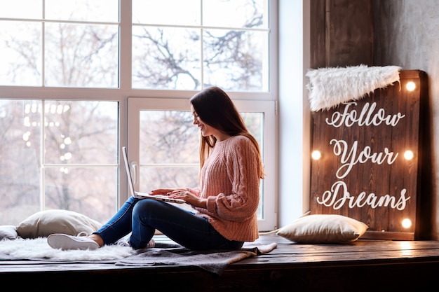 Mujer alegre está escribiendo en su computadora portátil mientras se queda en casa en auto aislamiento. Trabaja o estudia en línea.