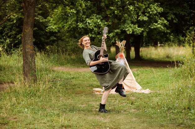 Mujer alegre está tocando la guitarra al aire libre en verano. La mujer rubia lleva vestido de lino y botas altas.