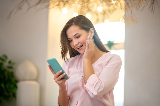 Mujer alegre feliz tocando su mejilla mientras está de pie con un teléfono inteligente en sus manos