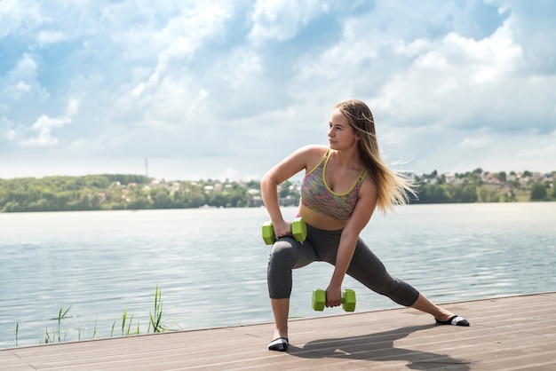 Mujer alegre fitness en ropa deportiva haciendo ejercicio con mancuernas, al aire libre. Estilo de vida saludable