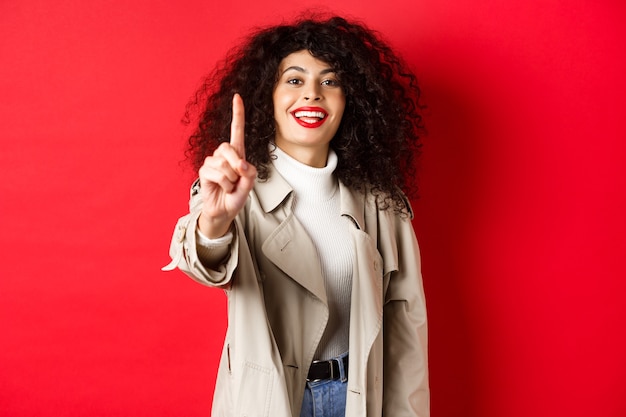 Mujer alegre en gabardina, mostrando el dedo número uno y sonriendo, de pie sobre la pared roja.