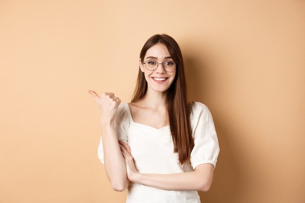 Mujer alegre en gafas apuntando con el dedo a la izquierda en el logo, sonriendo feliz, de pie sobre beige.