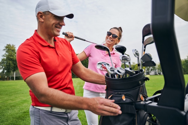 Mujer alegre en gafas de sol mirando a su instructor de golf poniendo su bolso en un coche