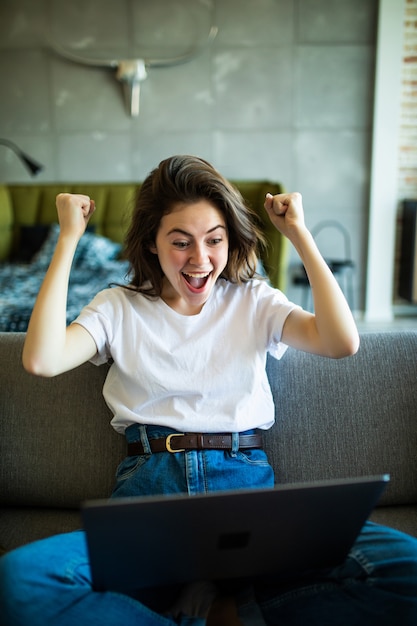 Foto mujer alegre con gesto de ganar usando la computadora portátil en el sofá