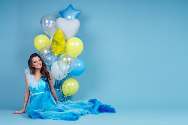 Mujer alegre con globos de helio azul, amarillo y blanco sentado y divirtiéndose de vacaciones en el estudio de fondo azul fiesta.Niña en un vestido largo posando maquillaje y peinado piel perfecta