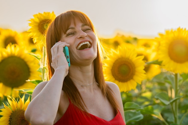 Mujer alegre hablando por su teléfono inteligente y disfrutando del día en un campo de girasoles.