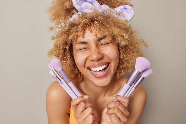 Una mujer alegre hace maquillaje decorativo, usa cepillos cosméticos, sonríe alegremente, tiene la piel limpia, usa una banda para la cabeza, mantiene los ojos cerrados, los hombros desnudos, aislados sobre un fondo gris, aplica cosméticos.