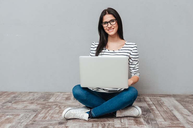 Foto mujer alegre joven con anteojos sentado en el piso mientras usa la computadora portátil. mirando al frente.