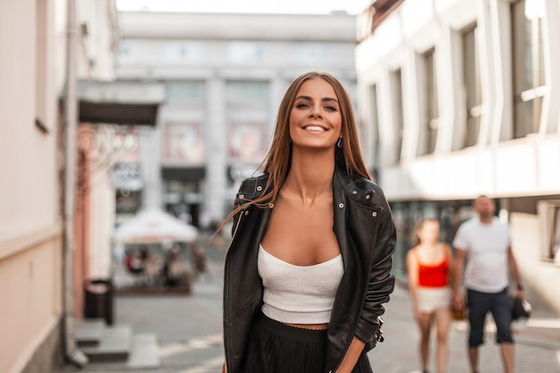 Mujer alegre joven inconformista con una sonrisa positiva en una blusa blanca de moda con una elegante chaqueta negra de cuero camina por la calle y disfruta de un soleado día de otoño. Chica alegre se relaja en la ciudad.