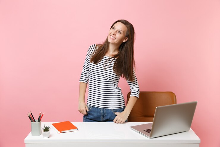Foto mujer alegre joven que mira a un lado el trabajo y que se coloca cerca del escritorio blanco con el ordenador portátil contemporáneo de la pc
