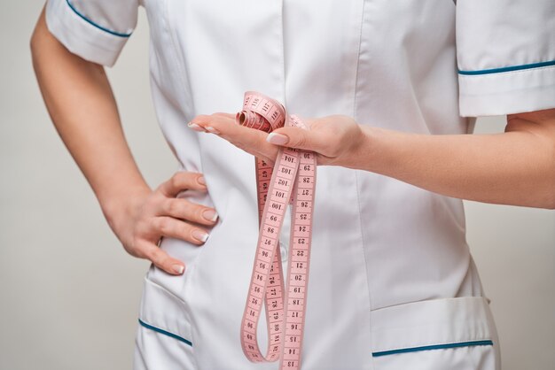 Mujer alegre médico o enfermera sosteniendo una cinta métrica de pie sobre una pared gris