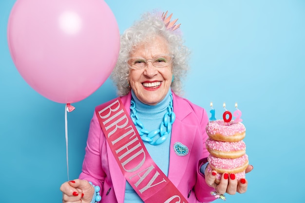 La mujer alegre y optimista celebra el cumpleaños número 102 sostiene una pila de donas y un globo de helio inflado vestido con ropa festiva se ve hermosa, bien cuidada, usa maquillaje brillante