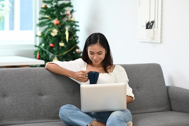 Una mujer alegre pasar el fin de semana en casa tomando café y usando la computadora portátil en el sofá.