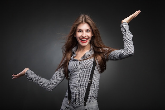 Mujer alegre posando con las manos en alto. Look de oficina con camisa a cuadros y tirantes. Retrato de estudio sobre fondo negro de viñeta.