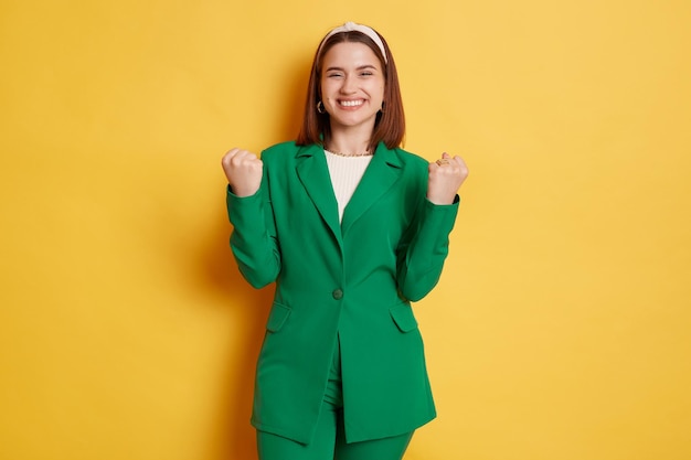 Una mujer alegre y positiva con una chaqueta verde que se encuentra aislada sobre un fondo amarillo con los puños apretados celebrando su victoria sonriendo con dientes