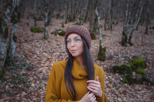 La mujer alegre positiva disfruta del otoño en el parque o el bosque usa suéter amarillo y sombrero, gafas. Estado de ánimo feliz chica hipster en viaje por carretera a finales de otoño por el bosque