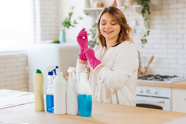 Mujer alegre positiva poniéndose guantes de goma mientras está lista para limpiar su casa