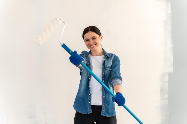 Mujer alegre y positiva sosteniendo y estirando a la cámara el rodillo sucio después de pintar las paredes de la habitación. Concepto de renovación y mejora del hogar de personas.