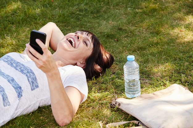 Mujer alegre que miente en hierba en parque con el teléfono