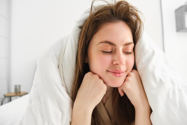 Foto mujer alegre que se siente cómoda en la cama acostada en el dormitorio cubierta con sábanas blancas sonriendo complacida