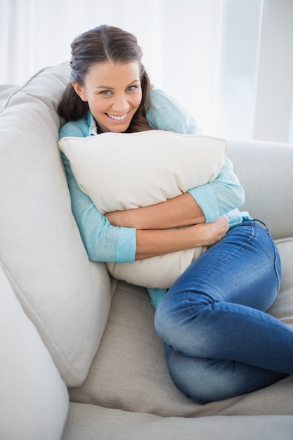 Mujer alegre que sostiene la almohada que se sienta en el sofá