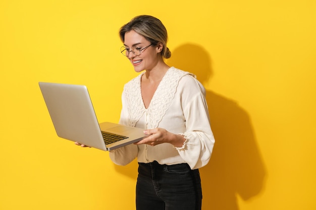 Una mujer alegre que usa anteojos está usando una computadora portátil con fondo amarillo