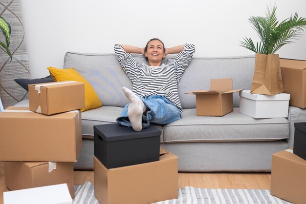 Mujer alegre reubicación se apoya en el sofá cajas desempaquetadas que significan el nuevo hogar feliz de bienes raíces