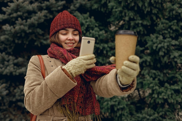 Mujer alegre en ropa de abrigo cálida tomando una foto de una taza de papel con café para llevar en el teléfono inteligente