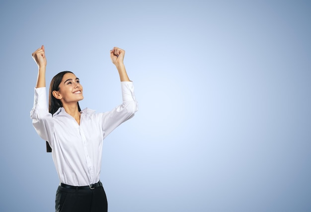 Mujer alegre y satisfecha con camisa blanca con las manos levantadas mostrando su ganador de poder y el concepto de logro aislado en un fondo azul claro con un lugar vacío para su maqueta de texto o logotipo