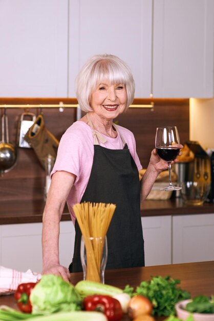 Mujer alegre senior está bebiendo vino tinto durante la cocción en la cocina moderna