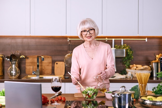 Mujer alegre senior está cocinando en la cocina moderna con un portátil