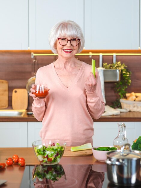 Mujer alegre senior con jugo de tomate y apio a dieta en su cocina