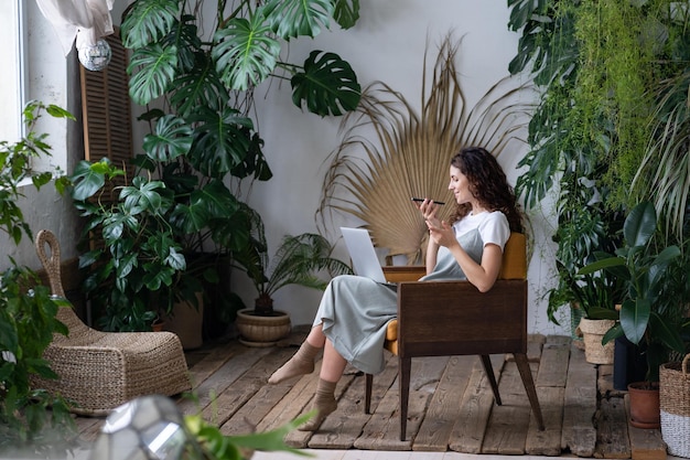 Foto mujer alegre sentada en el lugar de trabajo con una computadora portátil y grabando un mensaje de voz rodeada de plantas