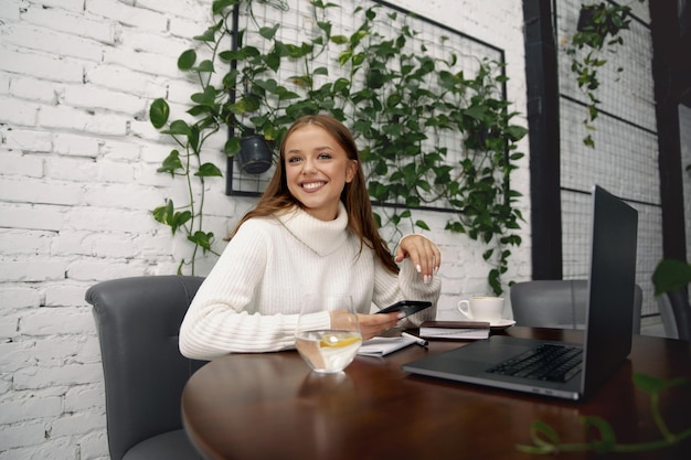 Mujer alegre sentada en la mesa estudiando usando una computadora portátil leyendo un libro