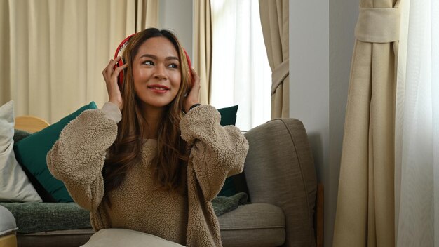 Mujer alegre sentada en la sala de estar y disfrutando escuchando música con auriculares inalámbricos