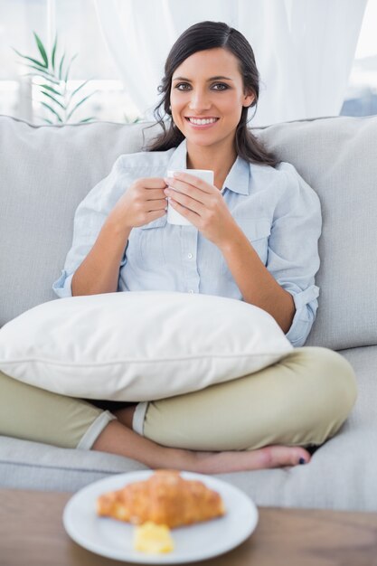 Mujer alegre en el sofá tomando café y teniendo croissant