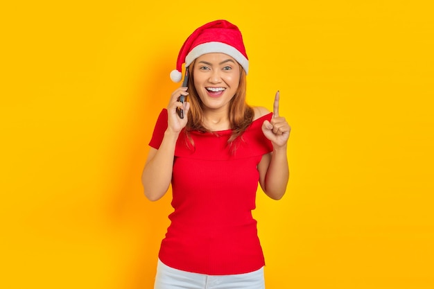 Mujer alegre con sombrero de Navidad hablando por teléfono móvil y apuntando con el dedo hacia arriba con una idea