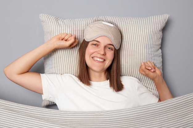 Mujer alegre y sonriente con camiseta blanca y antifaz para dormir tumbada en la cama sobre una almohada bajo una manta aislada en un fondo gris estirando las manos despertando