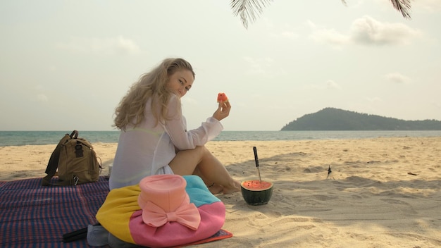 La mujer alegre sosteniendo y comiendo rebanadas de sandía en el mar de la playa de arena tropical