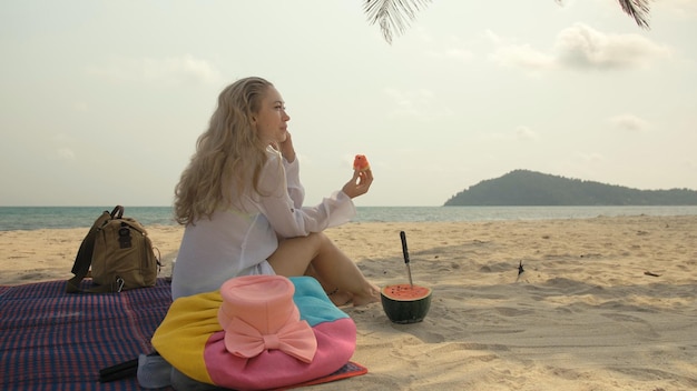 La mujer alegre sosteniendo y comiendo rebanadas de sandía en el mar de la playa de arena tropical