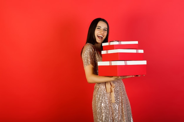 Mujer alegre sosteniendo una gran cantidad de cajas con regalos sobre un fondo rojo.