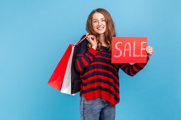 Mujer alegre con suéter de estilo casual a rayas que se jacta de compras con bolsas de papel y tarjetas de venta grandes descuentos de vacaciones Estudio interior aislado en fondo azul