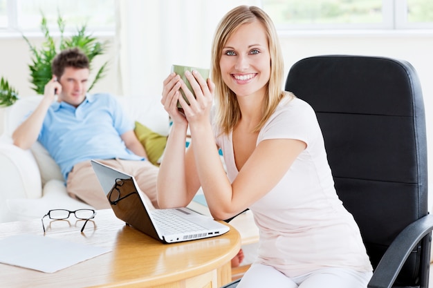 Mujer alegre con una taza de café y portátil con su novio sentado detrás de usar un teléfono celular