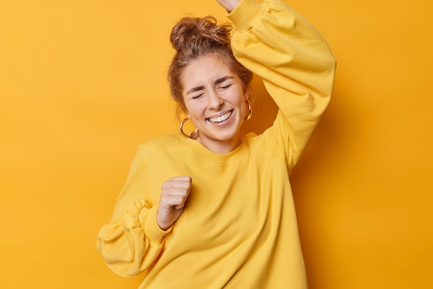 La mujer alegre tiene el pelo de buen humor recogido en un moño, levanta los brazos, baila con sonrisas de triunfo, usa aretes y un cómodo puente casual aislado sobre un fondo amarillo celebra los logros.