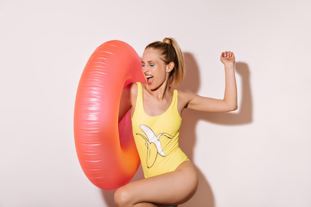 Mujer alegre en traje de baño de patrón de plátano amarillo regocijándose y posando con un gran anillo de natación rosa sobre fondo blanco