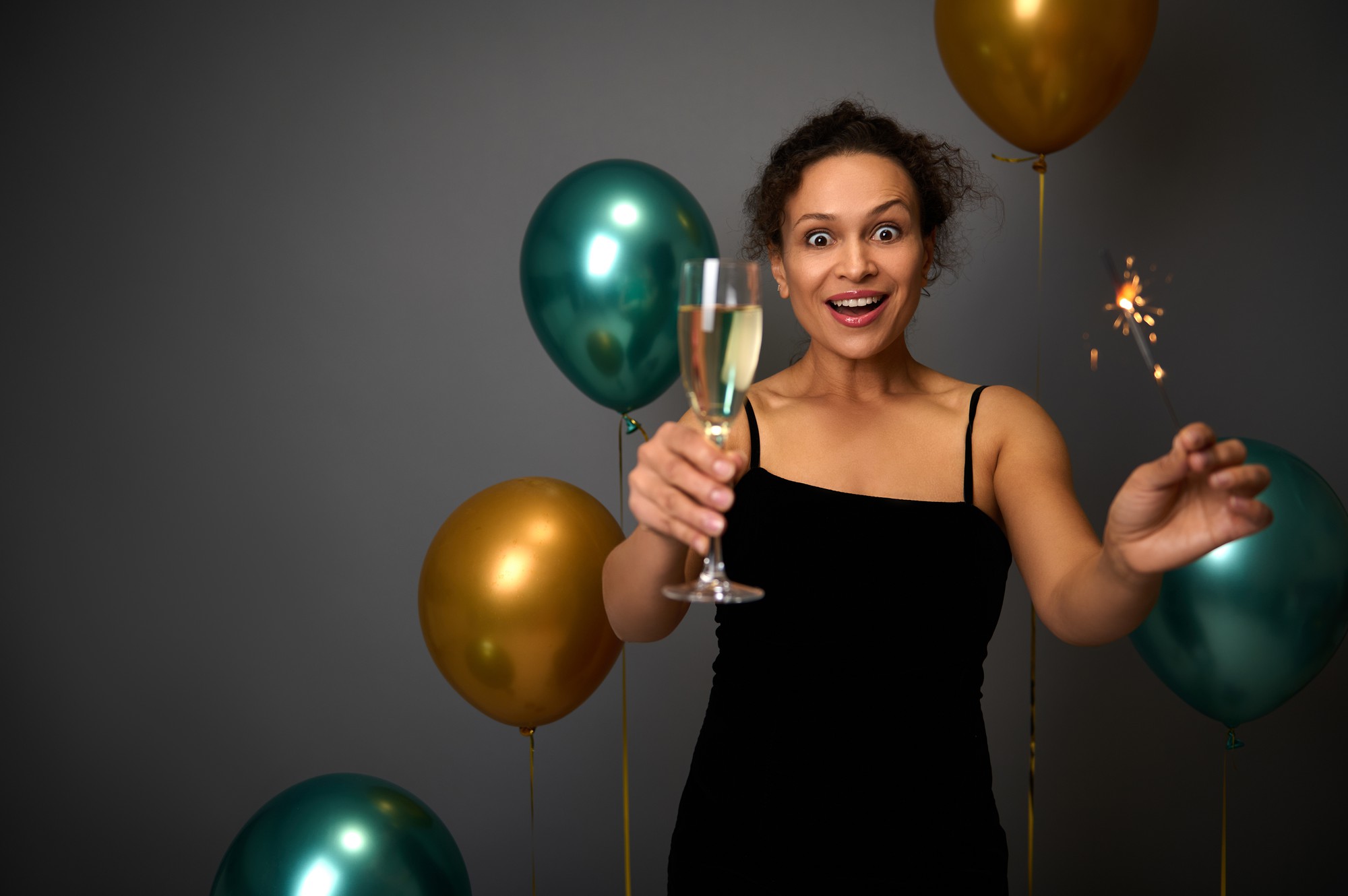 Mujer alegre en traje de noche negro sostiene una copa de champán y bengalas, posa sobre un fondo gris con una hermosa decoración de globos dorados y verdes. Navidad, concepto de cumpleaños