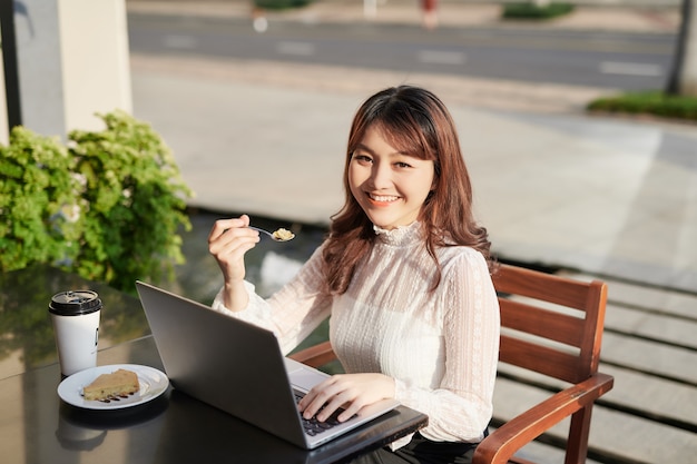 Mujer alegre usando laptop y comiendo pastel en un café.