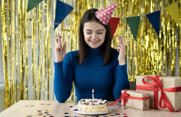 Una mujer alegre vestida con una gorra festiva en la cabeza se sienta en una mesa con un pastel y velas sonriendo alegremente Los dedos cruzados piden un deseo Fiesta en casa en línea Cumpleaños remoto