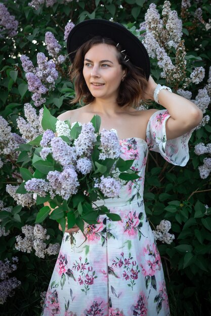 Una mujer alegre con un vestido ligero con hombros descubiertos y un estampado floral y un ramo de lilas.