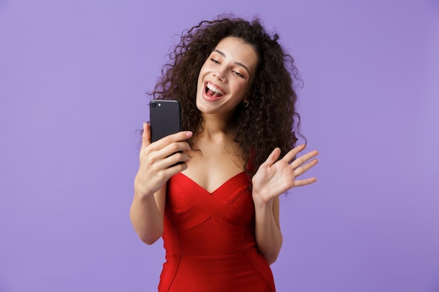 Mujer alegre con vestido rojo con smartphone negro, que se encuentran aisladas sobre la pared violeta